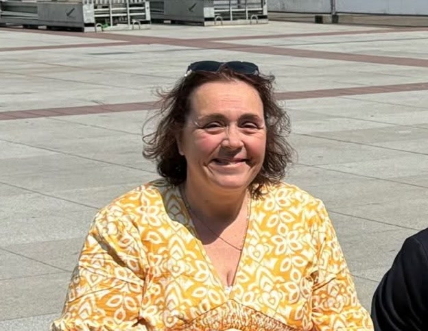 Ingrid Schoenmakers zit buiten op een zonnig plein en kijkt lachend naar de camera